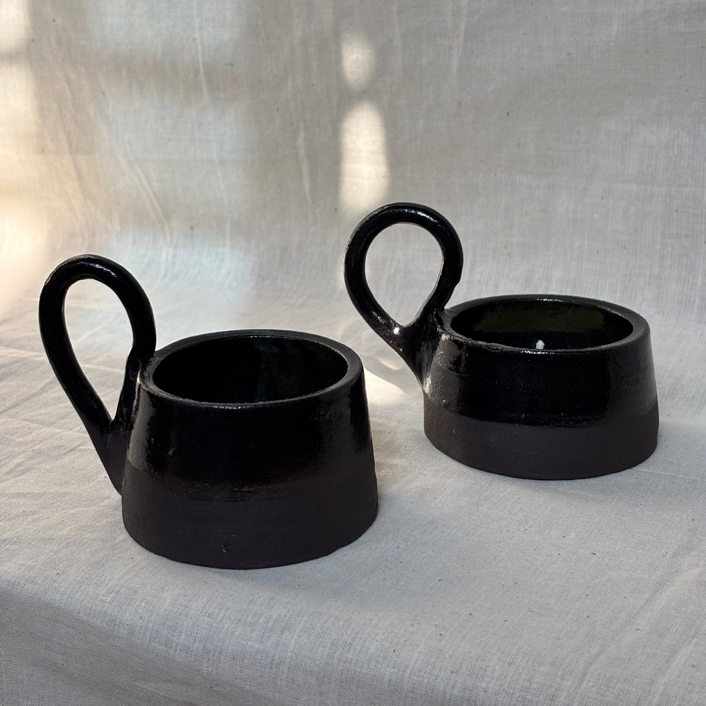 Two black ceramic candle holders with handles on a light fabric background