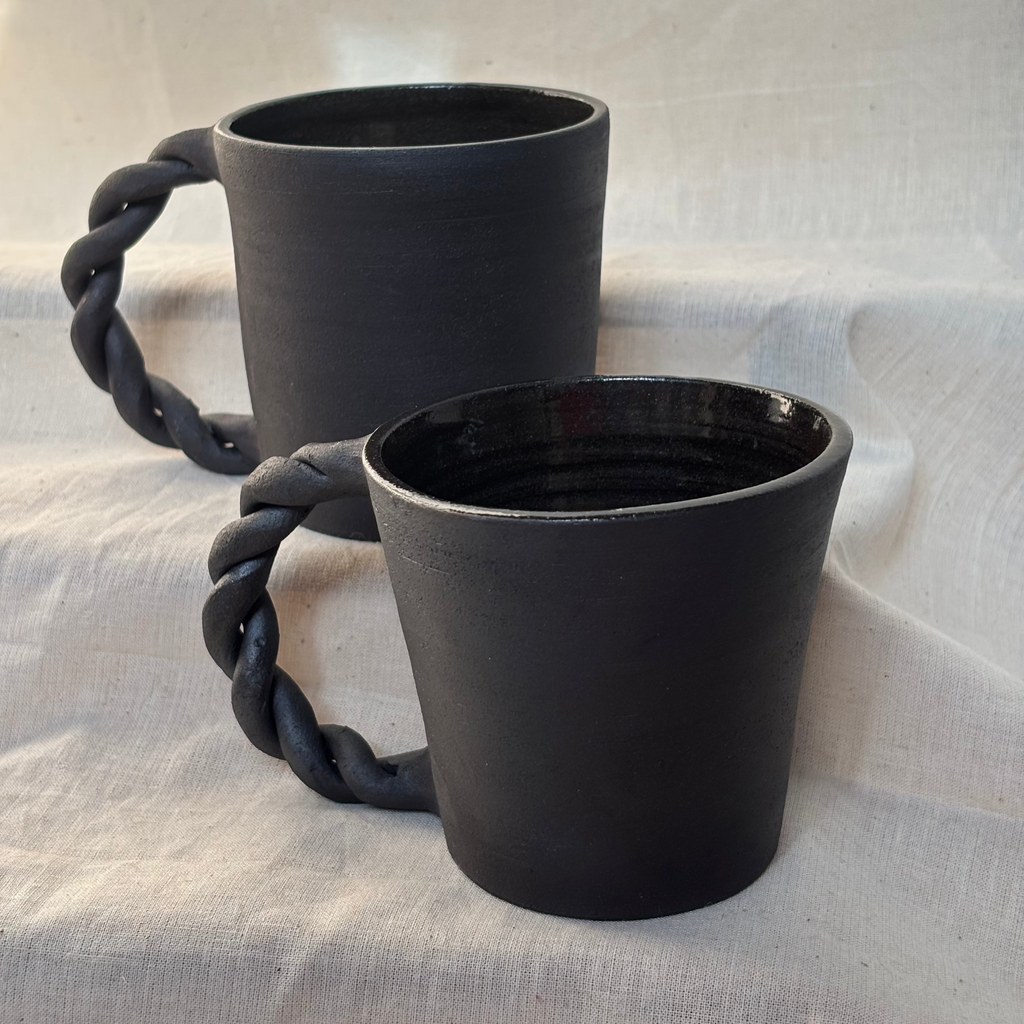 Two black ceramic mugs with rope handles on a light fabric background