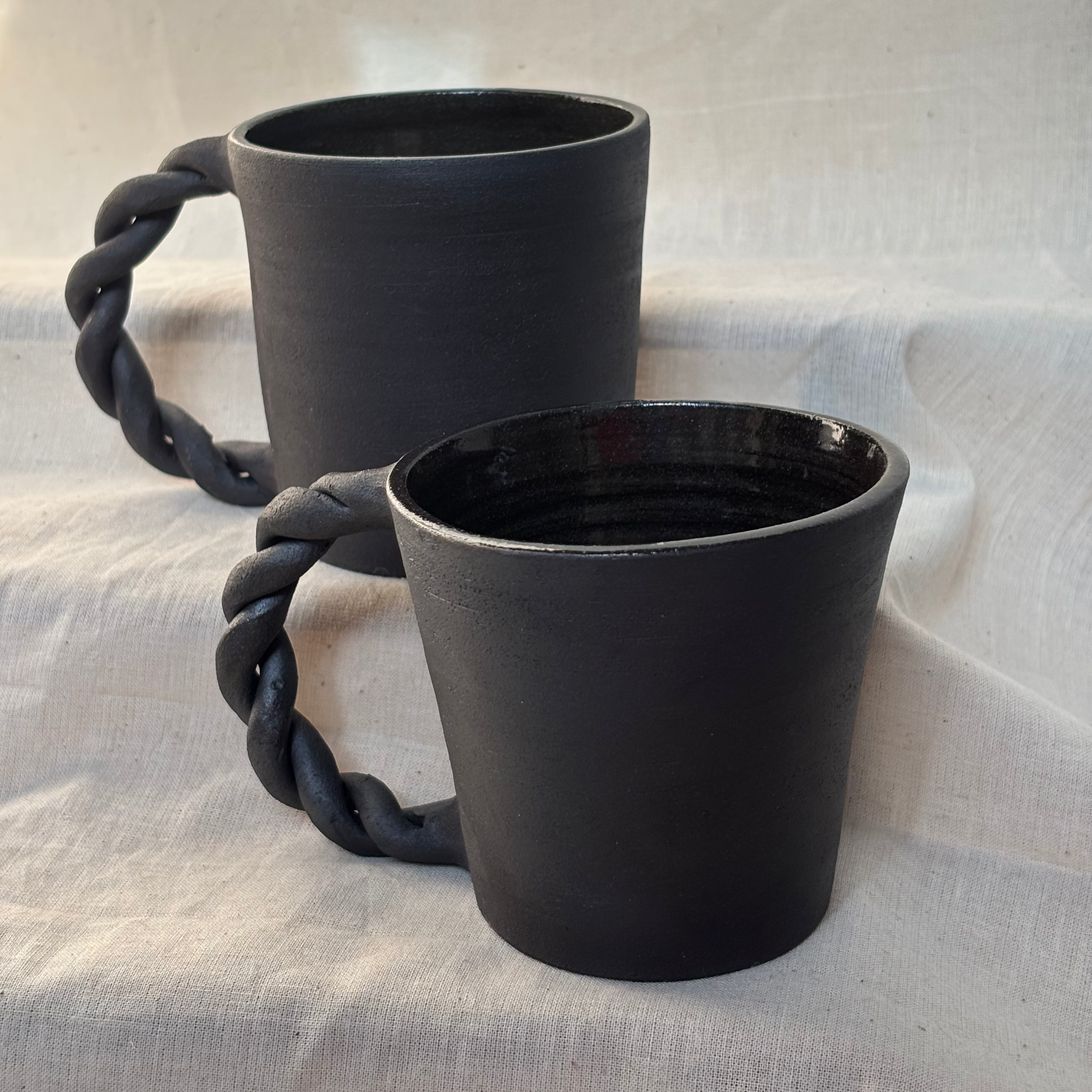 Two black ceramic mugs with rope handles on a light fabric background