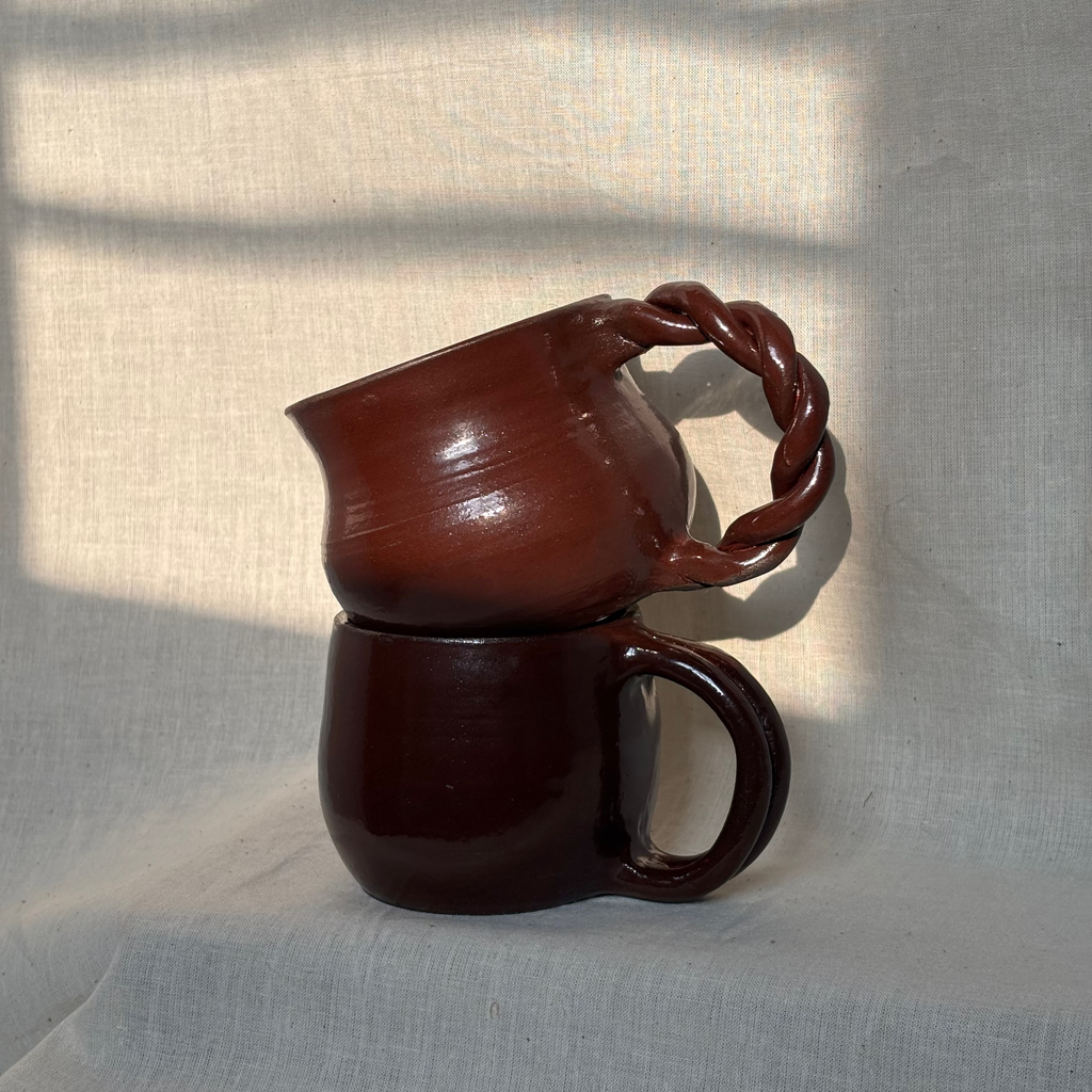 Two deep red ceramic mugs stacked on a textured light gray surface with shadows. One has a twisted handle and the other one has a smooth handle.