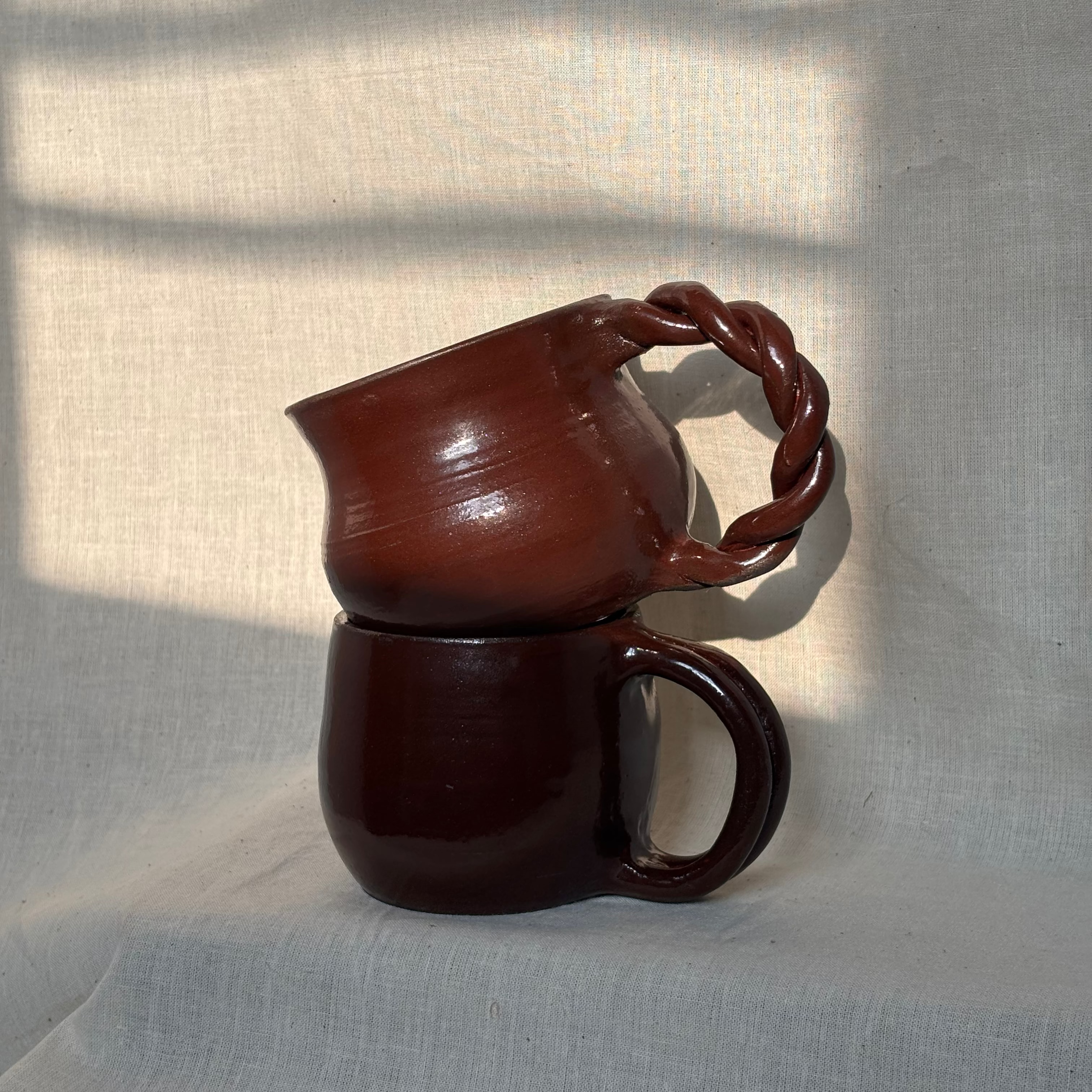 Two deep red ceramic mugs stacked on a textured light gray surface with shadows. One has a twisted handle and the other one has a smooth handle.